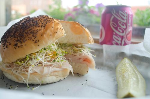 A poppy seed sandwich filled with sprouts and turkey, alongside a pickle and a can of Diet Coke.