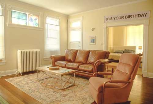 Cozy living room with a leather sofa, armchair, and a birthday banner on the wall, featuring warm lighting and wooden floors.