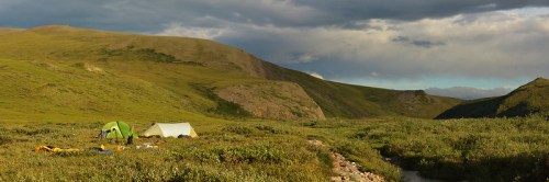A serene landscape featuring two tents set up in a lush green valley surrounded by rolling hills and cloudy skies.