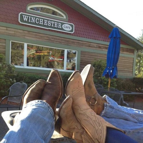 Two pairs of cowboy boots resting on a table in front of the Winchester Cafe, with a blue umbrella in the background.