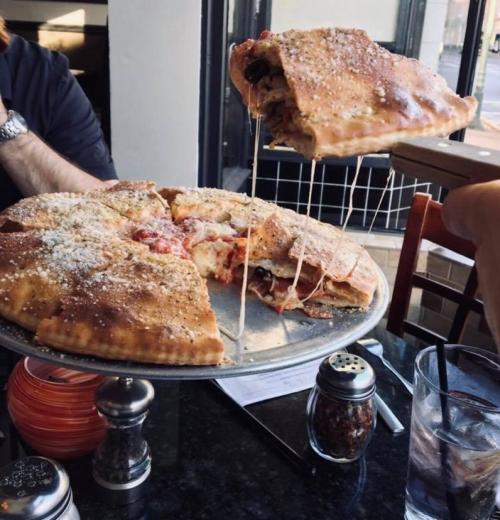 A large, cheesy calzone being lifted from a platter, with a slice revealing gooey cheese and toppings.
