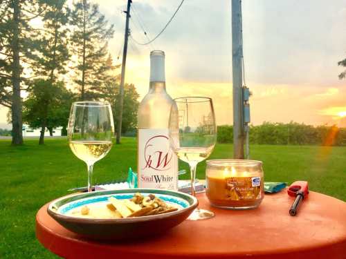 A table set with white wine, glasses, a plate of cheese and nuts, and a candle, with a sunset and trees in the background.