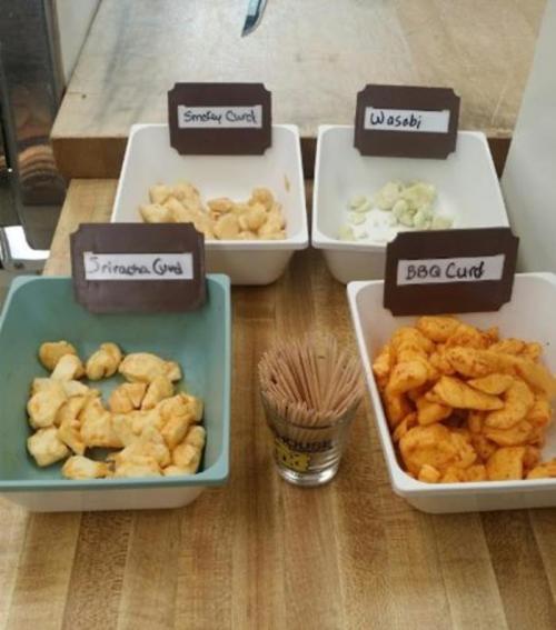Four bowls of flavored curds labeled Sriracha, Smoky, Wasabi, and BBQ, with toothpicks in a glass container.