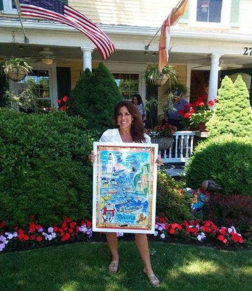 A woman holds a colorful artwork in front of a house with flowers and American flags.