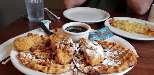 A plate of waffles topped with fried chicken, pecans, and powdered sugar, served with syrup on the side.