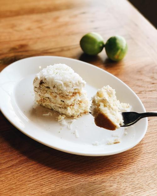 A slice of coconut cake on a white plate, with a fork and two limes in the background on a wooden table.