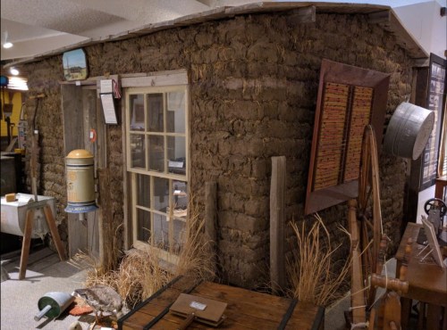 A rustic mud-brick building with a wooden door and window, surrounded by vintage tools and dry grass.