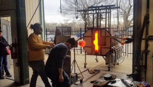 A glassblowing workshop with artists shaping molten glass in front of a furnace, surrounded by onlookers.