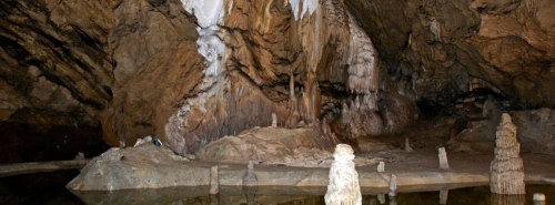 A dimly lit cave interior featuring stalactites, stalagmites, and a reflective pool of water.