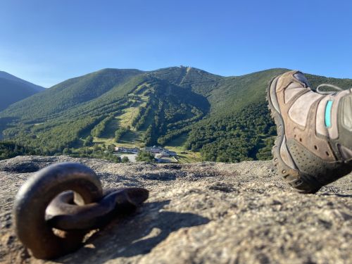 A hiker's boot rests on a rocky surface, overlooking a green mountain landscape and a ski resort below.