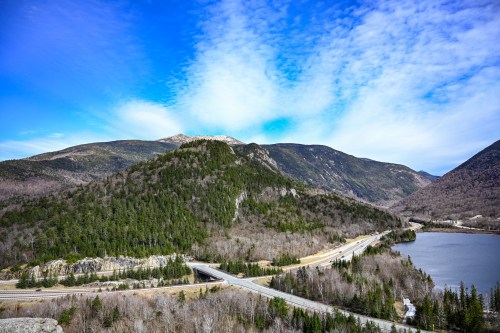 A scenic view of mountains and a lake under a blue sky, with a winding road in the foreground.