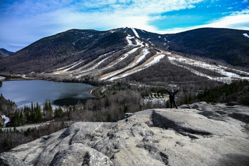 A person stands on a rocky outcrop overlooking a mountain landscape with ski trails and a lake below.