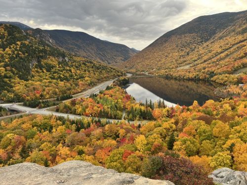 A scenic view of a valley with vibrant autumn foliage, a winding road, and a calm lake surrounded by mountains.