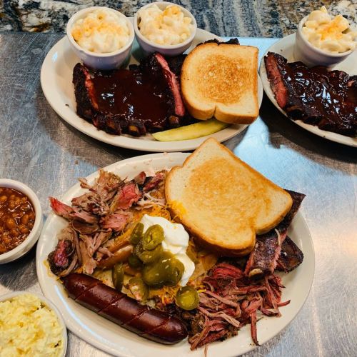 A spread of barbecue dishes including ribs, brisket, pulled pork, macaroni, baked beans, and toast on a table.