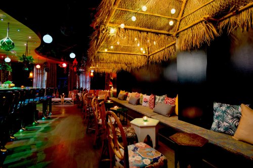 Cozy bar interior with tropical decor, bamboo seating, colorful cushions, and soft lighting.