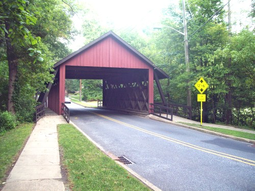 A red covered bridge spans a road, surrounded by green trees and a warning sign for low clearance.