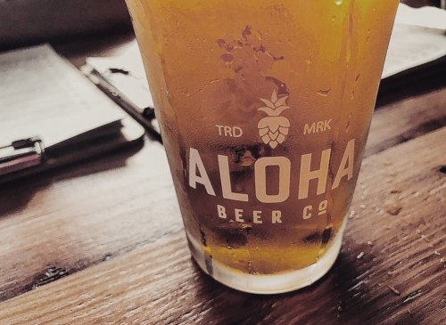 A close-up of a glass of beer with "Aloha Beer Co" logo on a wooden table, surrounded by menus and pens.