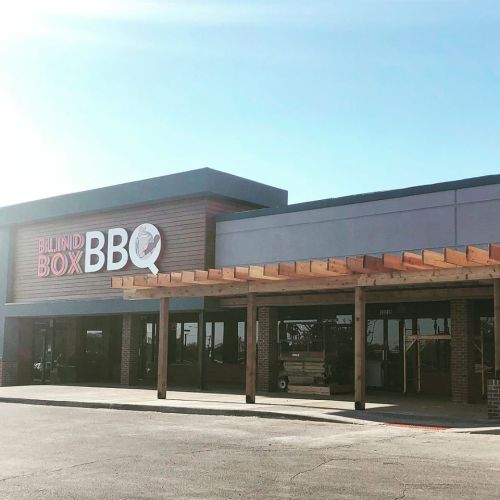 Exterior of Blind Box BBQ restaurant with a wooden awning and clear blue sky in the background.