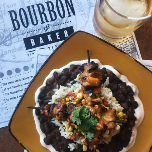 A plate of grilled chicken skewers on rice with black beans and corn, alongside a glass of bourbon.