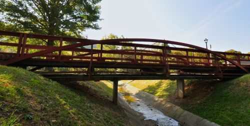 A red arched bridge spans a dry creek, surrounded by green grass and trees under a clear blue sky.