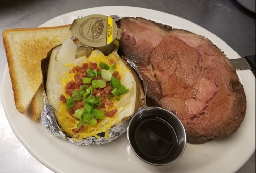 A plate featuring a slice of prime rib, a baked potato topped with cheese and green onions, toast, and a small cup of sauce.