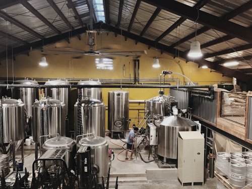 A brewery interior with large stainless steel tanks and a worker operating equipment in a spacious, industrial setting.