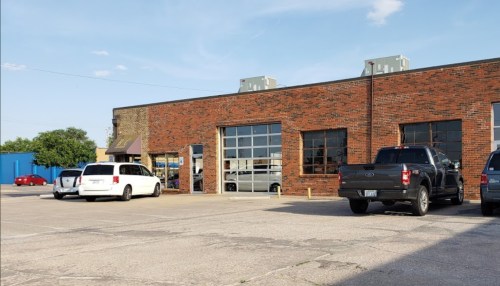A brick building with large windows, surrounded by parked cars in a paved lot under a clear blue sky.