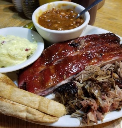 A plate of barbecue ribs, pulled pork, baked beans, and potato salad, with a slice of bread on the side.