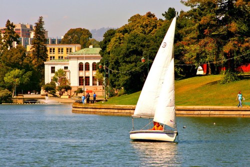 A sailboat glides across a calm lake, with trees and a building in the background on a sunny day.