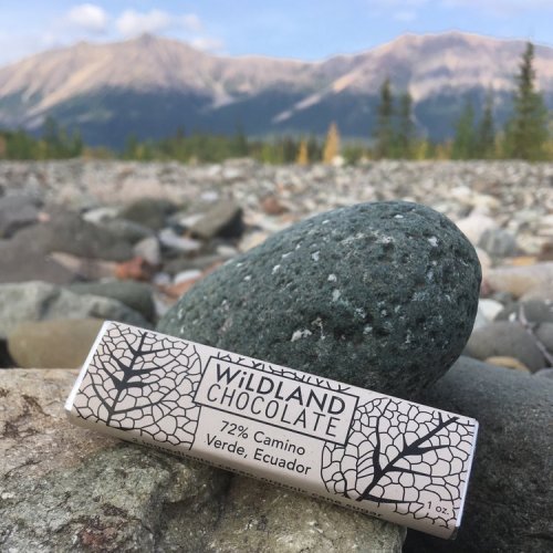 A chocolate bar from Wildland Chocolate rests on rocky terrain, with mountains and a blue sky in the background.