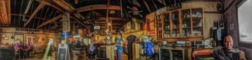 A cozy restaurant interior with wooden beams, patrons dining, and staff at the bar, creating a lively atmosphere.