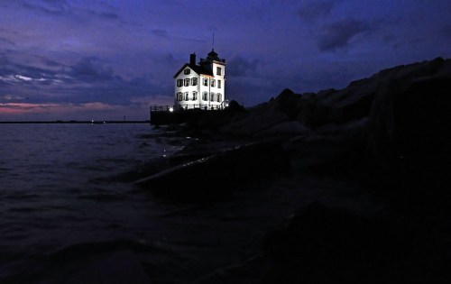 A lighthouse illuminated at dusk, standing on rocky shore with calm waters and a cloudy sky in the background.