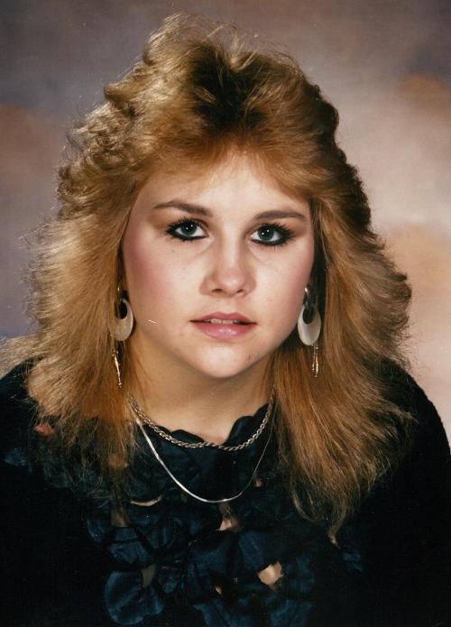 A young woman with voluminous blonde hair and bold makeup poses for a portrait, wearing large earrings and a black top.