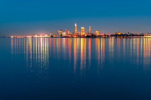 City skyline at dusk, reflecting lights on calm water, with tall buildings and a serene atmosphere.