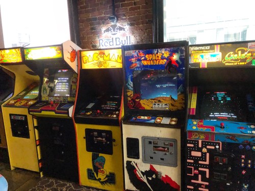 A row of vintage arcade game machines, including Pac-Man and Space Invaders, against a brick wall.