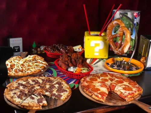 A table filled with various pizzas, chicken wings, and a pretzel, set against a colorful background.