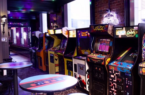 A row of vintage arcade machines in a dimly lit bar, with colorful lights and a brick wall backdrop.