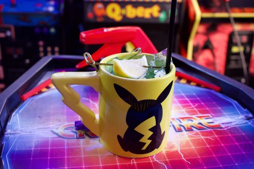 A vibrant yellow mug shaped like a character, filled with a cocktail, garnished with lemon and mint, on an arcade game table.