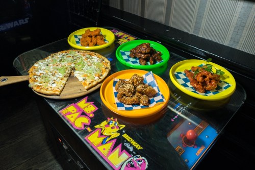 A wooden pizza paddle with a veggie pizza next to colorful plates of chicken wings and appetizers on an arcade game.