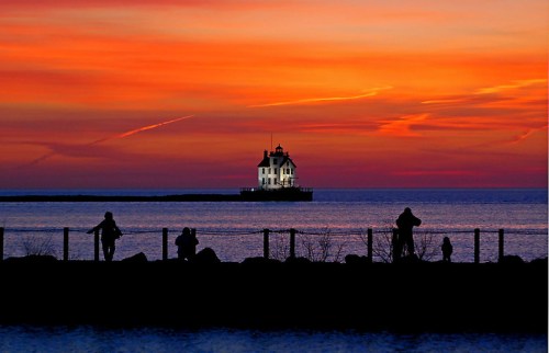 Silhouetted figures on a shore at sunset, with a lighthouse glowing against a vibrant orange and purple sky.