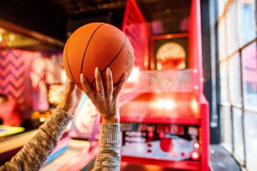 A person holds a basketball above their head in a vibrant arcade setting with a basketball game in the background.