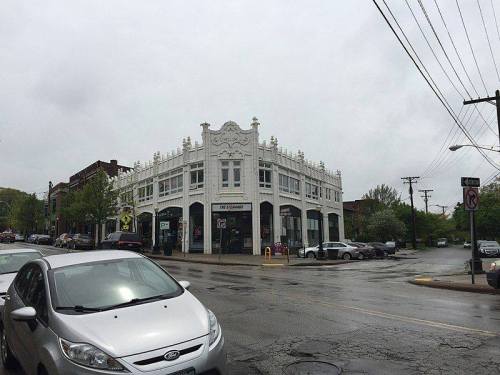 A white, ornate building at a street corner on a rainy day, with parked cars and trees in the background.