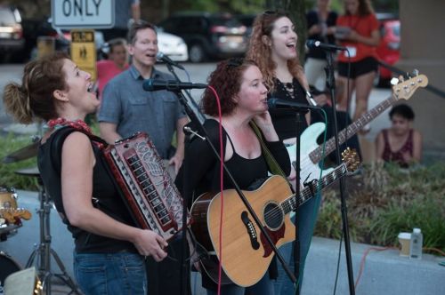 A lively band performs outdoors, featuring women with instruments and microphones, while an audience enjoys the music.