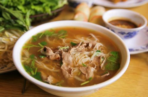 A bowl of noodle soup with beef, herbs, and green onions, served with fresh vegetables and dipping sauce on the side.