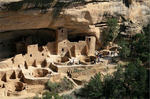 An aerial view of ancient cliff dwellings surrounded by trees, with visitors exploring the site.