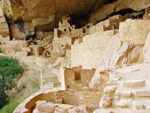 Ancient cliff dwellings built into a rocky overhang, surrounded by greenery and stone pathways.