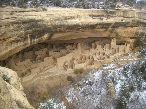 Ancient cliff dwellings nestled under a rocky overhang, surrounded by snow-covered trees and rugged terrain.