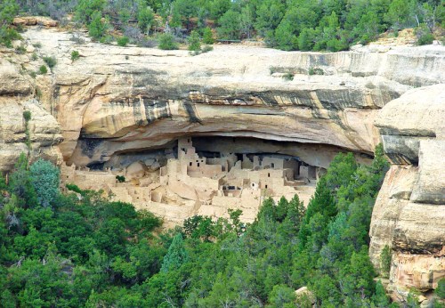Ancient cliff dwellings nestled under a rock overhang, surrounded by lush greenery and rugged terrain.