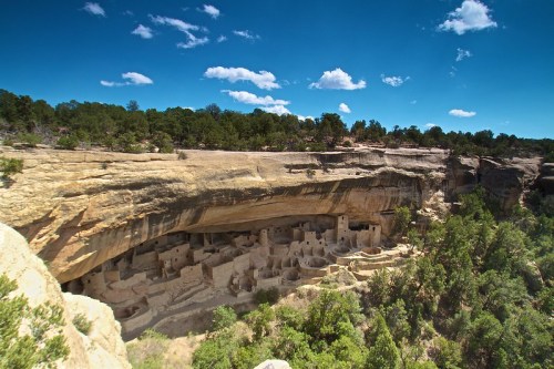 Ancient cliff dwellings nestled under a rocky overhang, surrounded by trees and a clear blue sky.