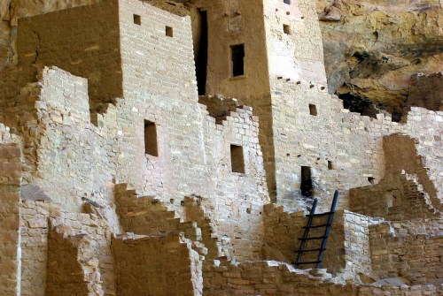 Ancient stone ruins of a cliff dwelling, featuring walls, windows, and a wooden ladder against a rocky backdrop.